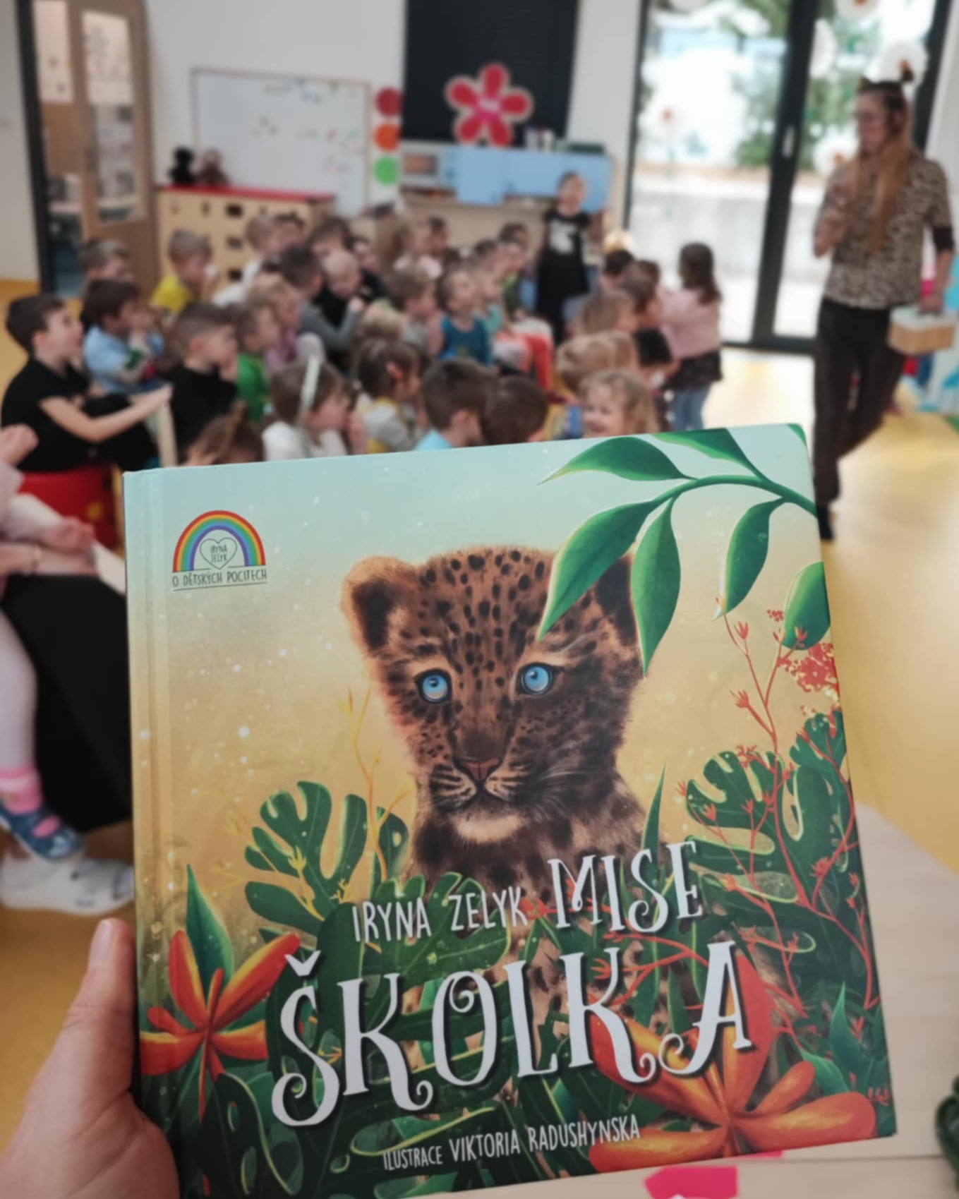 Fotka knížky Mise školka. V pozadí jde vidět herečku Marii a několik dětských diváků.